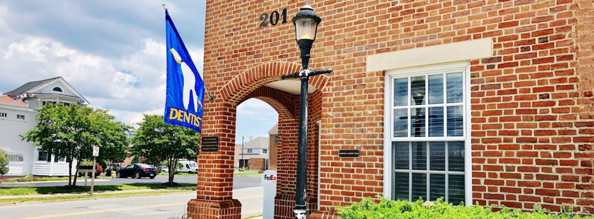The image shows a brick building with a sign that reads 101 above an arched doorway, which is adorned with a blue banner featuring a white horse logo. A flagpole stands in front of the building, displaying a blue and white flag. To the right of the entrance is a street lamp. The sky appears overcast, and there are trees visible behind the building.
