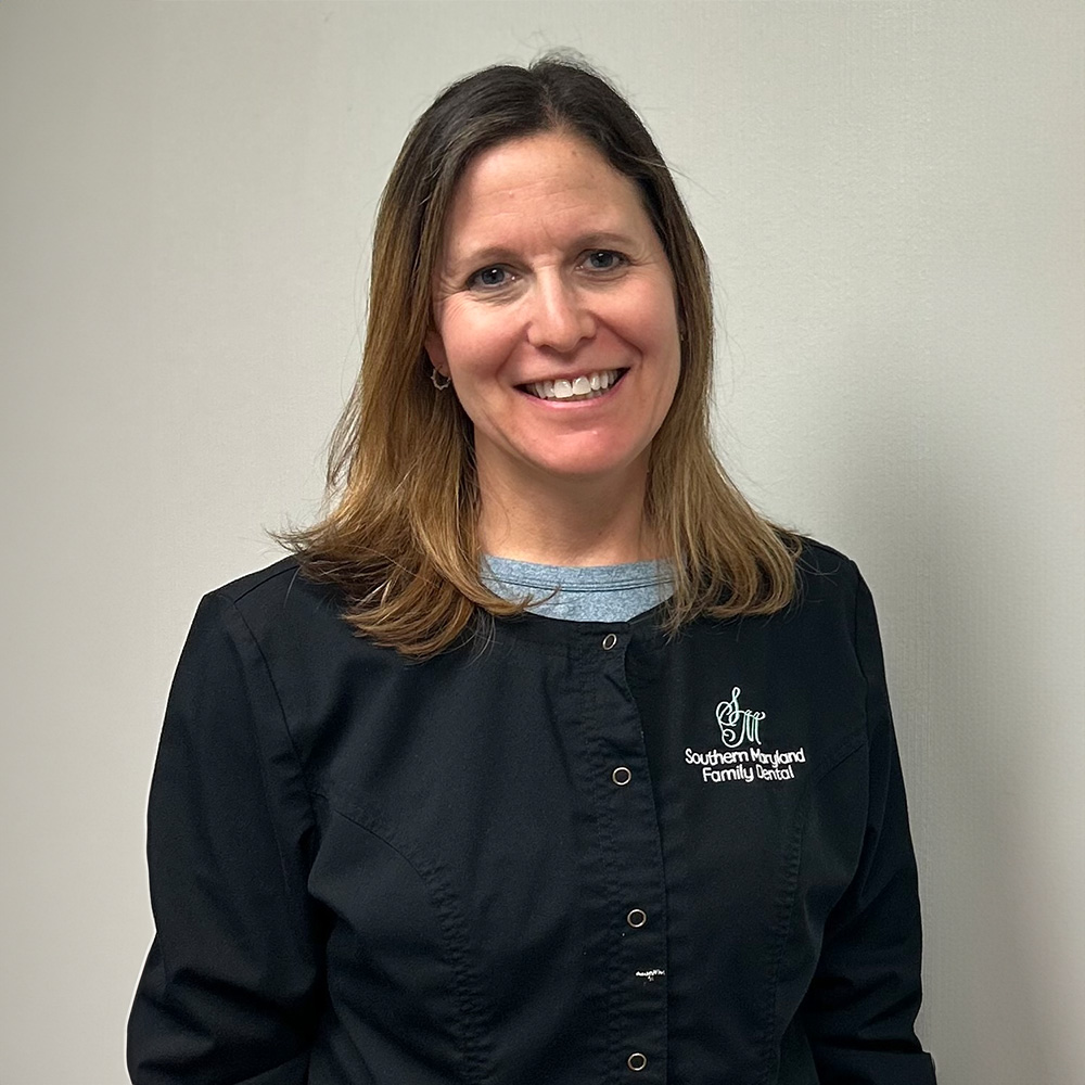 Woman standing indoors, wearing a black apron with a name tag, smiling at the camera.