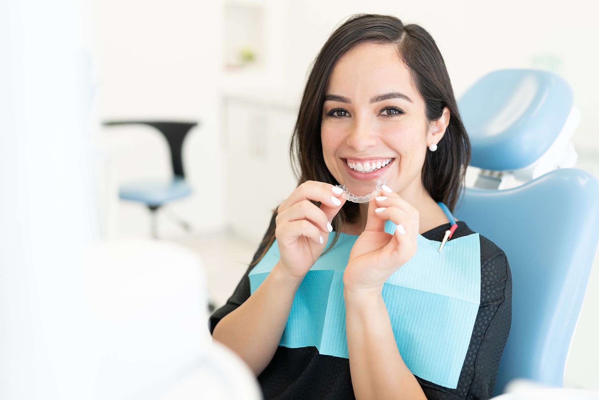 A woman is sitting in a dental chair, smiling, holding a toothbrush and looking at her teeth with a dental mirror in hand.